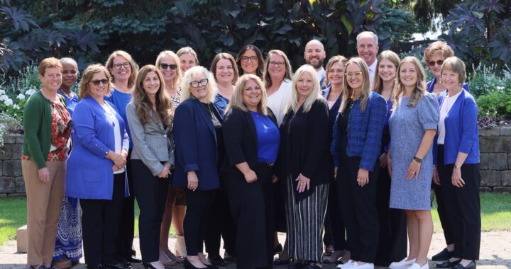 A group of 21 people, dressed in professional attire, poses outdoors on a sunny day. They are standing on a stone walkway with lush greenery and trees in the background. The group includes a mix of men and women, most of whom are wearing shades of blue, black, and white, with a few in other colors. Everyone is smiling, and they appear to be enjoying the moment together in a casual and friendly atmosphere.