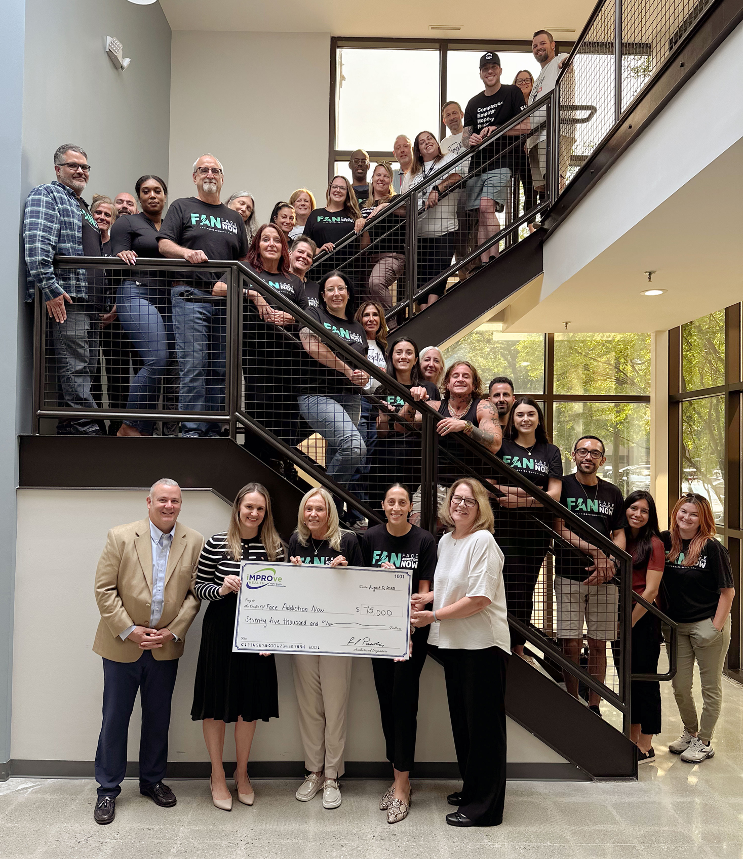 Members of the iMPROve Health staff present a check to FAN leadership with staff of FAN posed on a staircase behind them